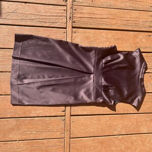 K&ouml;nnte beinhalten: Dunkelviolettes Satin-Kleid mit eng anliegendem Oberteil und plissiertem Rock. Das Kleid hat eine definierte Taille und einen dezenten Glanz, was auf einen formellen oder halbformellen Stil hindeutet. Das Kleid ist &auml;rmellos.