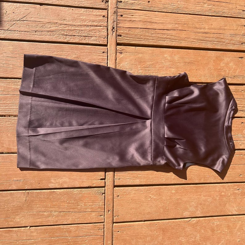 K&ouml;nnte beinhalten: Dunkelviolettes Satin-Kleid mit eng anliegendem Oberteil und plissiertem Rock. Das Kleid hat eine definierte Taille und einen dezenten Glanz, was auf einen formellen oder halbformellen Stil hindeutet. Das Kleid ist &auml;rmellos.