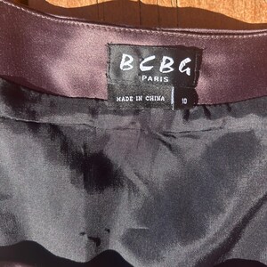 K&ouml;nnte beinhalten: Nahaufnahme eines dunkelvioletten Satin-Kleidungsst&uuml;cks. Das Etikett lautet "BCBG PARIS" und "MADE IN CHINA". Das Gr&ouml;&szlig;enetikett zeigt Gr&ouml;&szlig;e 10 an. Der Stoff scheint ein dunkles, gl&auml;nzendes Material zu sein.