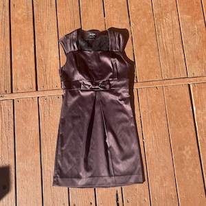 K&ouml;nnte beinhalten: Ein pflaumenfarbenes Satinkleid mit Schleife in der Taille. Das Kleid hat einen plissierten Rock und Fl&uuml;gel&auml;rmel. Der Ausschnitt hat eine quadratische Form mit einem schwarzen Netzeinsatz. Das Kleid ist mini-lang.