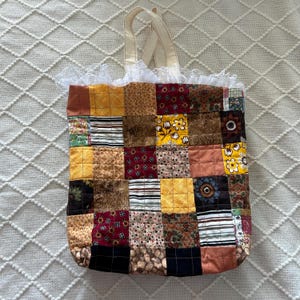 May include: A patchwork tote bag with a variety of fabric squares in shades of brown, yellow, and red. The bag has cream-colored handles and a white lace trim along the top edge. The bag is made of quilted fabric.