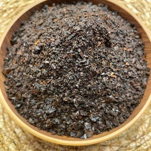 May include: A wooden bowl filled with dark brown, coarse flakes. The flakes have a slightly reddish hue and a textured appearance. The bowl sits on a woven, light brown surface, creating a natural, earthy aesthetic.