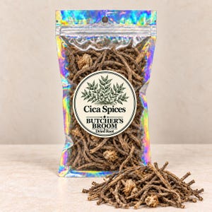 Può includere: Una busta olografica riempita con radici essiccate di pungitopo marroni. Un'etichetta circolare sul davanti presenta un'illustrazione botanica e il testo Cica Spices, Butcher's Broom e Dried Root. Un piccolo mucchio di radici è posto davanti.