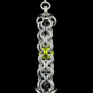 May include: A vertical chainmail pendant featuring interlocking silver, black, and lime green metal rings. The pendant is suspended from a thin black cord and displays a complex woven pattern with a single row of bright lime green rings.