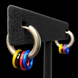 May include: A pair of silver-toned hoop earrings, each featuring three small, smooth rings in vibrant yellow, black, red, and blue colors suspended from the main hoop. The earrings are displayed on a black T-shaped stand against a dark background.