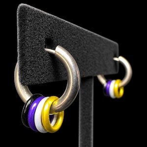 Puede incluir: Un par de pendientes de aro en tono plateado, cada uno con tres pequeños anillos lisos en colores negro, morado, blanco y amarillo ensartados en el aro principal. Los pendientes se muestran en un soporte en forma de T negro sobre un fondo negro.
