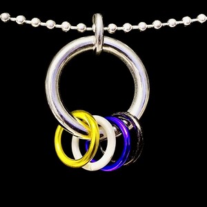 Peut inclure: Un collier en métal argenté avec un grand pendentif en forme d'anneau. Le pendentif contient quatre petits anneaux jaunes, blancs, bleus et noirs. La chaîne du collier est composée de petites perles argentées reliées, sur fond noir.