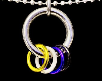 Könnte beinhalten: Halskette aus silberfarbenem Metall mit einem großen Ringanhänger. Der Anhänger hält vier kleinere Ringe in Gelb, Weiß, Blau und Schwarz. Die Halskette besteht aus kleinen, verbundenen Silberperlen, vor schwarzem Hintergrund.
