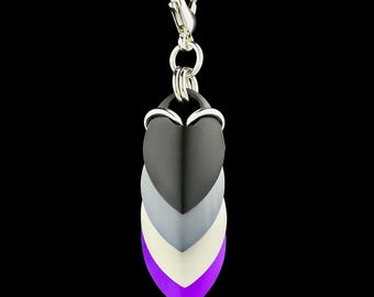 Könnte beinhalten: Eine silberne Halskette mit einem Anhänger mit einem geschichteten Herzdesign in Schwarz, Grau, Weiß und Lila. Der Anhänger ist an einem silbernen Verschluss befestigt. Der Hintergrund ist schwarz.