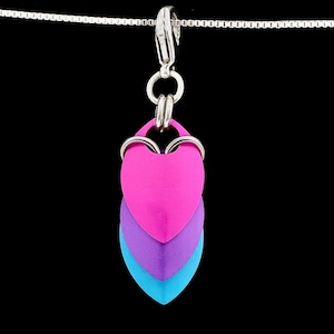 Op de afbeelding: Zilveren ketting met een hanger van drie hartvormige metalen schubben in roze, paars en blauw. De hanger is met een zilveren sluiting en ringen aan de ketting bevestigd, tegen een zwarte achtergrond.