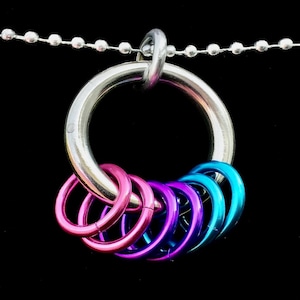 Könnte beinhalten: Ein silberfarbener Metallring-Anhänger mit sechs kleineren, farbigen Ringen. Die kleineren Ringe sind rosa, lila und blau. Der Anhänger hängt an einer silbernen Kugelkette vor schwarzem Hintergrund.