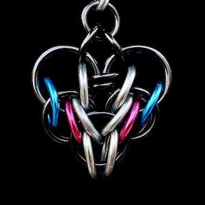 Op de afbeelding: Een zilverkleurige maliënkolder hanger met een complex, verweven ontwerp. De hanger heeft een hartvorm met blauwe en roze accenten. De achtergrond is effen zwart, wat het ingewikkelde metaalwerk benadrukt.