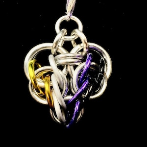 Pode incluir: Um pingente de malha de corrente feito à mão com um design de bandeira de orgulho não binário. O pingente é feito de anéis de metal prateado, com detalhes em anéis dourados, brancos, roxos e pretos. Ele pende de um cordão roxo e branco contra um fundo preto.