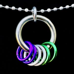 May include: A silver-colored metal pendant necklace featuring a large ring with smaller, colorful rings attached. The smaller rings are purple, white, and green. The necklace chain is made of small, silver-colored beads.