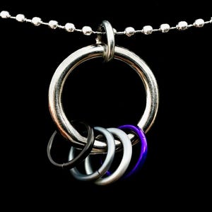 Op de afbeelding: Zilverkleurige ketting met een grote ringhanger en vijf kleinere ringen in zwart, zilver, lichtblauw en paars. De ketting heeft een kogelketting en een zwarte achtergrond.