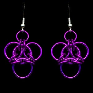Puede incluir: Un par de pendientes de cota de malla morados hechos a mano. Cada pendiente presenta un diseño floral con anillos metálicos entrelazados. Los pendientes tienen ganchos plateados y un acabado metálico brillante. El fondo es negro liso.