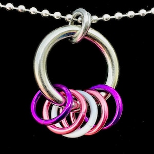 Op de afbeelding: Zilverkleurige ketting met een grote, zilveren ringhanger. De hanger heeft een reeks kleinere ringen in roze, paarse en witte tinten. De ketting wordt gepresenteerd tegen een zwarte achtergrond.