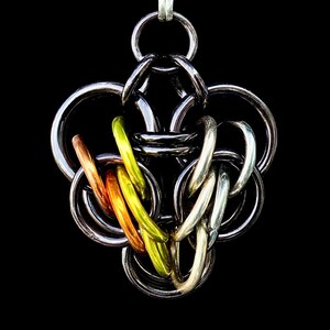 Op de afbeelding: Een handgemaakte hanger met een complex kanten ontwerp. De hanger is voornamelijk zilverkleurig, met accenten van koper, oranje en geelgroen. Het ingewikkelde metaalwerk is geplaatst tegen een zwarte achtergrond.