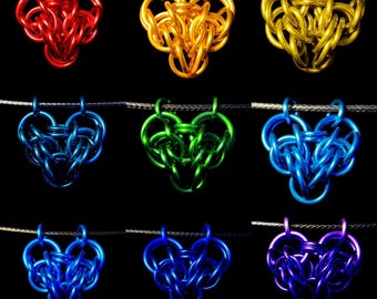 Puede incluir: Nueve colgantes en forma de corazón en varios colores, incluyendo rojo, dorado, amarillo, azul, verde y morado. Cada colgante está hecho de anillos metálicos entrelazados, suspendidos de un fino cordón sobre un fondo negro.