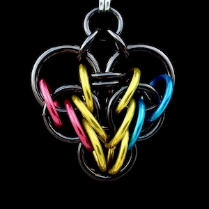 Op de afbeelding: Een hangerketting met een complex kanten ontwerp. De ketting heeft een mix van zilveren, gouden, roze en blauwe ringen, waardoor een kleurrijk en ingewikkeld patroon ontstaat. De achtergrond is effen zwart.