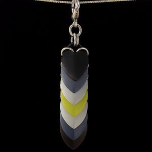 Op de afbeelding: Een hangerketting met hartvormige bedels in een kleurengradatie. De bedels zijn zwart, grijs, wit en limoengroen, en hangen aan een zilveren ketting. De achtergrond is effen zwart.