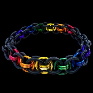 Puede incluir: Una pulsera de cota de malla negra con anillos metálicos de colores del arcoíris. La pulsera presenta una serie de anillos entrelazados en un espectro de colores, incluyendo rojo, naranja, amarillo, verde, azul y morado. Fondo negro.