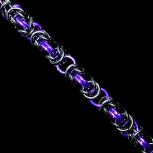 Op de afbeelding: Een close-up van een maliënkolderketting. De ketting heeft een patroon van in elkaar grijpende ringen in zilver, zwart en helder paars. De ketting wordt weergegeven tegen een zwarte achtergrond, waardoor de metallic tinten worden benadrukt.