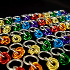 Puede incluir: Primer plano de una colección de anillos de cota de malla coloridos. Cada anillo es de un color diferente, incluyendo rojo, naranja, amarillo, verde, azul y morado. Los anillos están conectados con anillas plateadas, creando un patrón texturizado.