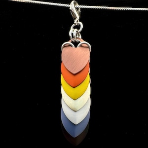 Op de afbeelding: Een kleurrijke hangerketting met hartvormige metalen schubben in koper, oranje, geel, crème, wit en blauw. De hanger hangt aan een zilveren ketting en sluiting, tegen een zwarte achtergrond.