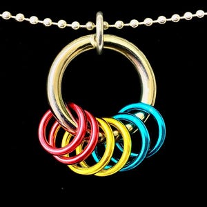 Op de afbeelding: Een goudkleurige metalen ringhanger met een ketting, met vijf kleinere ringen in rood, geel en blauw. De hanger hangt tegen een zwarte achtergrond en benadrukt de heldere kleuren van de ringen.