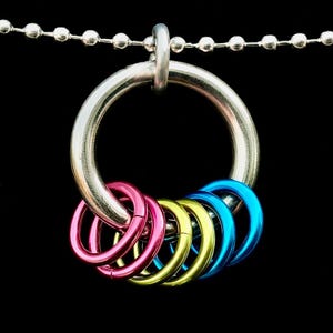 Op de afbeelding: Een zilverkleurige metalen ringhanger met zes kleinere, kleurrijke ringen eraan. De kleinere ringen zijn roze, geel en blauw. De hanger hangt aan een zilveren kogelketting tegen een zwarte achtergrond.