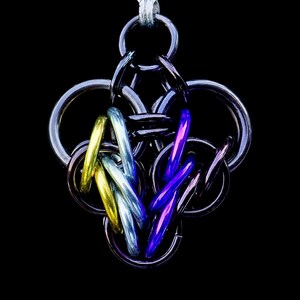 Könnte beinhalten: Eine handgefertigte Kettenhemd-Anhänger-Halskette. Der Anhänger hat ein komplexes Design aus ineinandergreifenden Metallringen in Silber-, Gold-, Blau- und Lilatönen. Die Halskette hängt vor einem schwarzen Hintergrund, der die komplizierten Details und Farben hervorhebt.