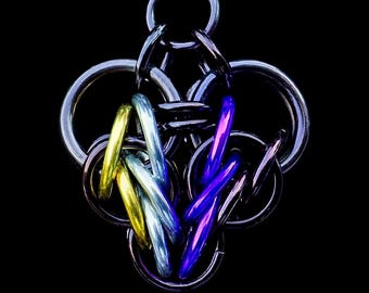 Könnte beinhalten: Eine handgefertigte Kettenhemd-Anhänger-Halskette. Der Anhänger hat ein komplexes Design aus ineinandergreifenden Metallringen in Silber-, Gold-, Blau- und Lilatönen. Die Halskette hängt vor einem schwarzen Hintergrund, der die komplizierten Details und Farben hervorhebt.