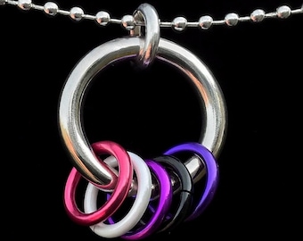 May include: A silver-colored necklace featuring a large ring pendant with six smaller, colorful rings attached. The smaller rings are pink, white, purple, and black. The necklace has a ball chain.