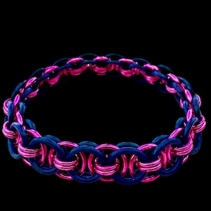 Può includere: Un braccialetto fatto a mano con un design a maglia di catena. Il braccialetto è composto da anelli ad incastro nei colori rosa acceso e blu scuro. Il braccialetto è mostrato su uno sfondo nero.
