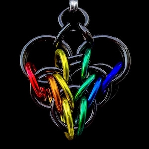 Op de afbeelding: Een handgemaakte chainmaille hanger in hartvorm. De hanger is gemaakt van zilverkleurige ringen, met gekleurde regenboogringen erdoorheen geweven. De achtergrond is effen zwart.