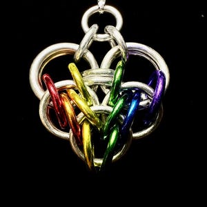 Op de afbeelding: Een zilverkleurige hangerketting met een hartvormig ontwerp. De hanger heeft verweven metalen ringen en kleurrijke, regenboogkleurige accenten. De achtergrond is effen zwart, waardoor de ingewikkelde details van de hanger worden benadrukt.