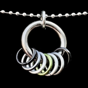 Op de afbeelding: Een zilverkleurige ketting met een grote, cirkelvormige hanger en verschillende kleinere ringen in diverse metallic tinten. De ketting heeft een kogelketting en een zwarte achtergrond.