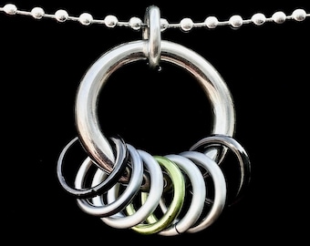 May include: A silver-colored necklace featuring a large, circular pendant with a series of smaller rings in various metallic hues. The necklace has a ball chain and a black background.