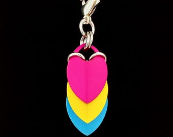 Könnte beinhalten: Eine Halskette mit Anhänger, die einen Charm mit drei herzförmigen Schuppen in Pink, Gelb und Blau aufweist, die an einer Silberkette hängen. Der Charm ist an einem Silberverschluss befestigt. Der Hintergrund ist schwarz.