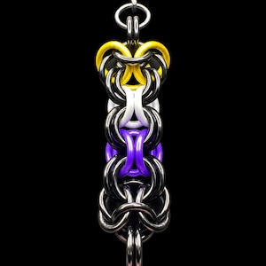 May include: A chainmail pendant featuring a vertical arrangement of interlocking rings in yellow, white, purple, and black. The pendant is suspended from a silver-toned metal bail and a textured gray cord, showcasing a complex woven metal pattern.
