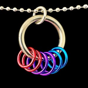 Op de afbeelding: Een zilverkleurige metalen sleutelhanger met een grote ring en een ketting van kleine zilveren balletjes. Aan de grote ring zijn zes kleinere ringen in rood, paars en blauw bevestigd. De achtergrond is effen zwart.