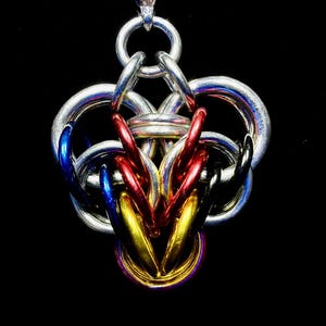 May include: A handcrafted chainmaille pendant with a complex, interwoven design. The pendant features silver, red, blue, black, and gold-colored rings, creating a striking visual contrast against the black background. The intricate metalwork suggests a unique, artistic piece.