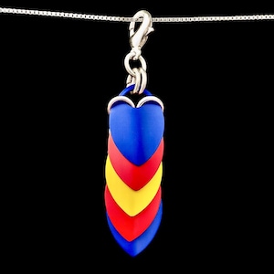 Op de afbeelding: Een ketting met hanger, bestaande uit een ketting en een bedel gemaakt van gelaagde hartvormige stukken in blauw, rood en geel. De bedel hangt aan een zilverkleurige sluiting. De achtergrond is zwart.