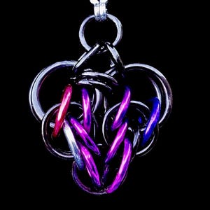 May include: A silver-colored chainmail pendant with a complex, interwoven design. The pendant features a mix of silver, purple, red, and blue rings, creating a striking contrast against the black background. The pendant is suspended from a silver chain.