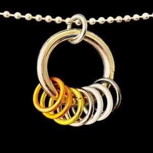Op de afbeelding: Een zilverkleurige metalen ring met een reeks kleinere ringen in goud-, brons- en zilverkleurige tinten. De grote ring hangt aan een kogelketting, alles tegen een zwarte achtergrond. Het sieraad is een hanger.