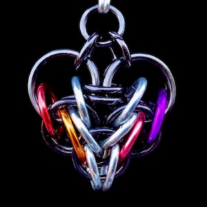 Op de afbeelding: Handgemaakte hanger met een complex kanten ontwerp. De hanger is voornamelijk zilverkleurig, met accenten van rode, oranje, roze en paarse ringen. Het ingewikkelde weefsel vormt een hartvorm, opgehangen aan een zilveren lus tegen een zwarte achtergrond.