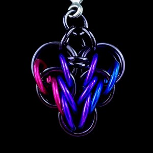 Op de afbeelding: Een hangerketting met een complex ontwerp van in elkaar grijpende metalen ringen. De hanger bevat roze, blauwe en paarse ringen, met een zilveren ketting. De achtergrond is effen zwart.