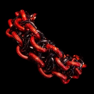 Peut inclure: Gros plan sur un bracelet composé de maillons métalliques rouges et noirs entrelacés. Le bracelet est posé sur un fond noir, soulignant le contraste des couleurs et le design complexe de la cotte de mailles.