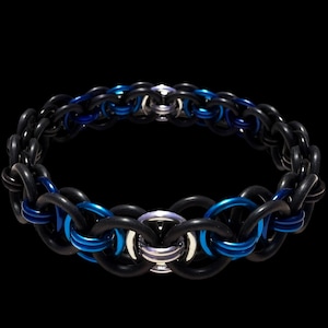 May include: A chainmail bracelet featuring a pattern of black and blue rings. The bracelet has a central silver-colored clasp. The links are made of metal and are arranged in an intricate pattern. The background is black.
